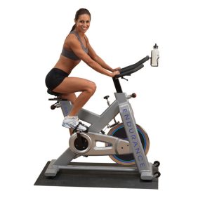 A woman is sitting on a Body Solid Endurance Indoor Bike - New in a gym equipped with new and remanufactured gym equipment.