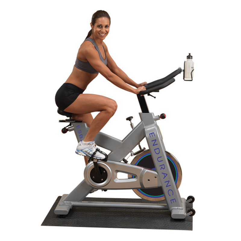 A woman is sitting on a Body Solid Endurance Indoor Bike - New in a gym equipped with new and remanufactured gym equipment.
