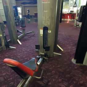 The Freemotion Calf - As Is Functional machine features a red and black seat, elegantly placed on a maroon patterned carpet. Behind it, a collection of modern fitness equipment stands ready, offering a complete workout experience.
