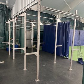The Rig - Remanufactured metal workout frame includes pull-up bars and various attachments, making it ideal for your indoor gym. Complemented by dark flooring and blue divider curtains, it offers a versatile training experience.