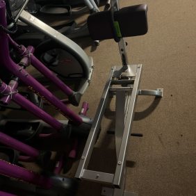 A remanufactured Cybex Bent Leg Abdominal Board, featuring a padded seat and support handles, sits on a beige carpeted floor, nestled among other fitness equipment.
