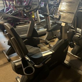 In a dimly lit indoor setting, rows of stationary exercise bikes align with precision, some boasting screens partially covered in plastic. Experience the elevated workout environment with the Life Cycle - Remanufactured bike from Fitness Plus, where every pedal stroke brings you closer to your fitness goals.