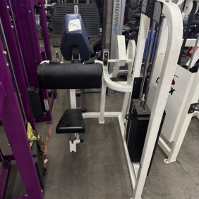 The ProMaxima Plate Loaded Lat Pulldown - As Is Functional comes with a black cushioned seat and backrest, surrounded by other gym equipment, providing a comprehensive workout experience.