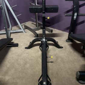 A serviced gym setup features a black metal frame with adjustable components and yellow knobs, including the Hoist Olympic Decline Bench - Serviced, all set on a carpeted floor against a striking purple wall.