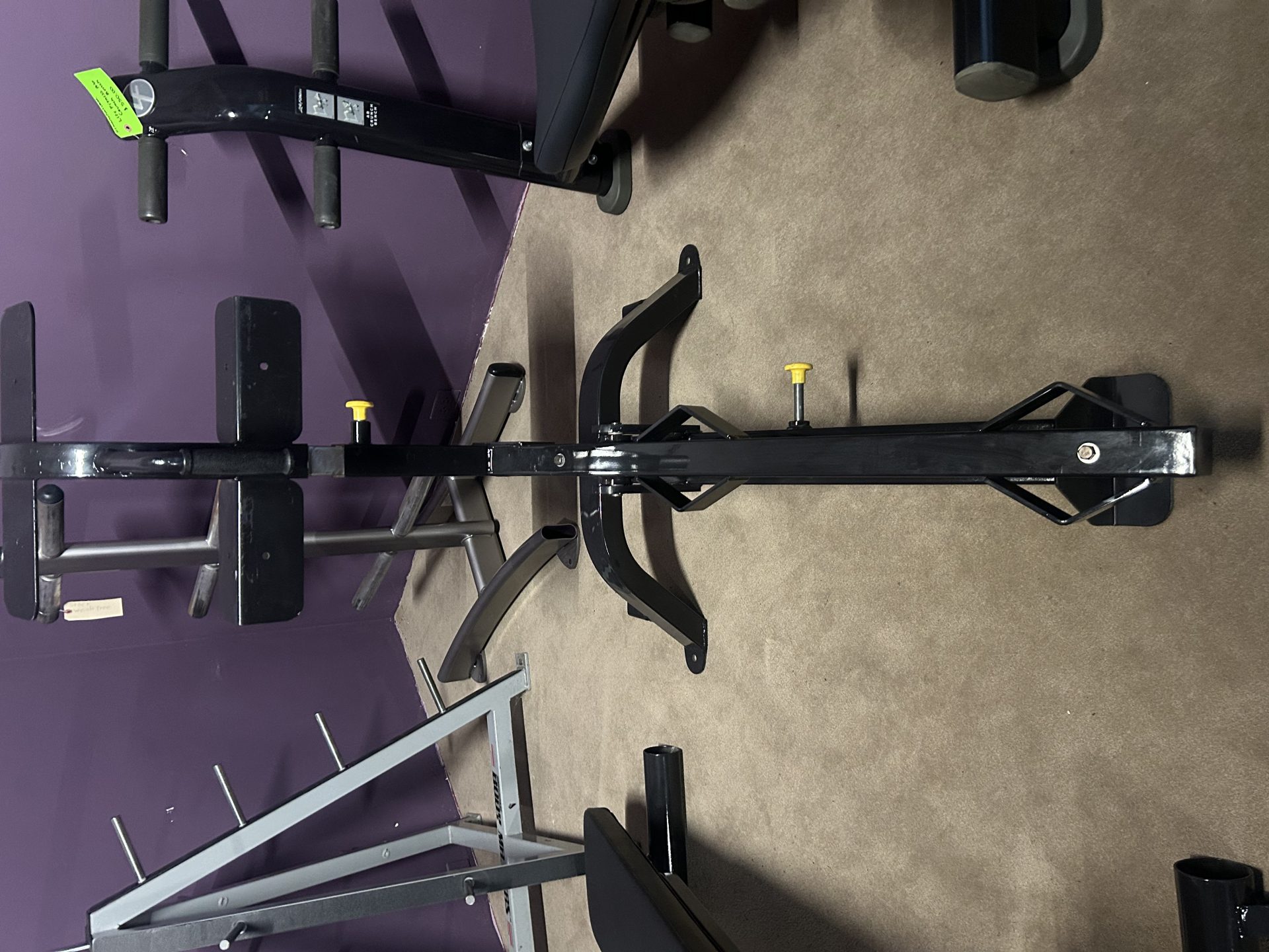 A serviced gym setup features a black metal frame with adjustable components and yellow knobs, including the Hoist Olympic Decline Bench - Serviced, all set on a carpeted floor against a striking purple wall.