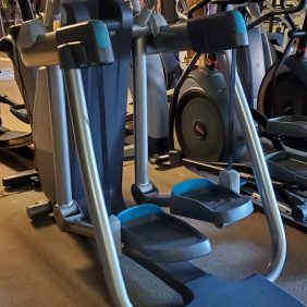 In a gym setting, two elliptical machines are positioned side by side, including a serviced Precor AMT835, surrounded by other fitness equipment.