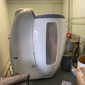 A white cryotherapy chamber labeled "extraCRYO" is positioned in a utility room with pipes, cleaning supplies, and a trash can nearby.