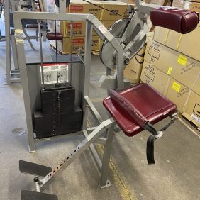 A Cybex Back Extension - As Is Functional with red padding and adjustable weights is set up in a warehouse, located near stacked cardboard boxes.