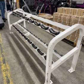 A white dumbbell rack stands in a busy warehouse filled with boxes and gear, ready for use. This inviting space encourages workouts, hinting at the Matrix Leg Press - As Is Functional nearby to meet fitness enthusiasts' needs.