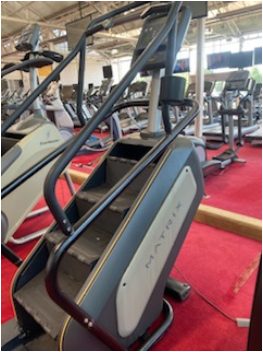 A remanufactured Matrix Climbmill ($3,295) sits on a red carpeted gym floor, surrounded by treadmills and ellipticals.