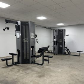 Two Cybex strength training machines are placed in a spacious gym room with plain white walls and a tiled ceiling. The floor is bare concrete.