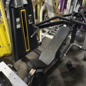 A seated chest press machine with padded backrest and handles is positioned among other gym equipment in a fitness center.