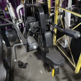 A seated gym machine for chest and shoulder exercises with padded arms, handles, and a foot lever, surrounded by other fitness equipment.