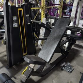 A commercial chest press exercise machine with a black padded seat and backrest is positioned in a warehouse among other gym equipment.