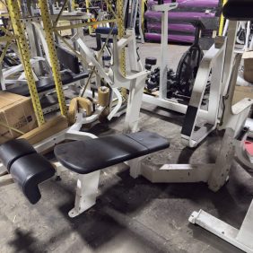 A gym storage area with white Freemotion Epic Olympic Decline Bench - As Is Functional, along with other benches and machines, on a concrete floor beneath yellow metal shelving.