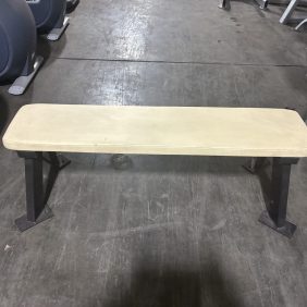 A flat weight bench with a beige padded top and metal legs, positioned on a gym floor surrounded by fitness equipment.