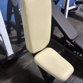 A close-up view of a beige padded seat and backrest on a black metal exercise machine, set on a concrete floor next to a blue wall.