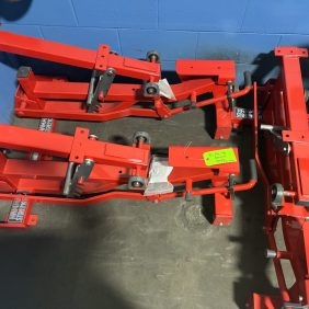 Three red hydraulic floor jacks, one with a green label, sit on a concrete floor against a blue wall beside a remanufactured Hammer Strength Flat - 90 Degree Bench.