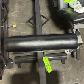 Black padded gym equipment with worn foot pads and green tags attached, positioned on a concrete floor near other exercise machines.
