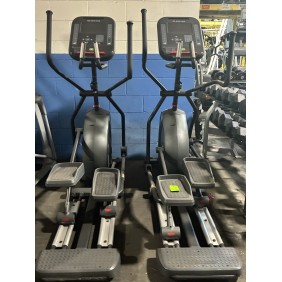 Two elliptical exercise machines are positioned side by side in a gym with a blue and white painted wall in the background.