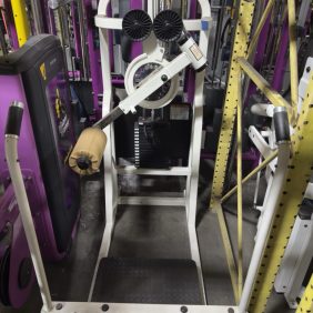 A standing calf raise machine with padded shoulder rests, a foot platform, and weight stack, located in a gym with purple and yellow equipment.