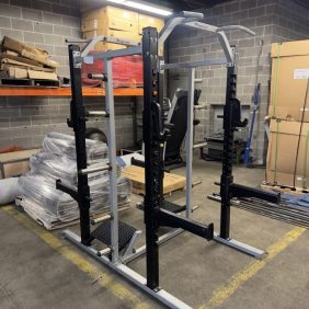 A squat rack with adjustable safety bars is positioned in a storage area with gym equipment, pallets, and boxes against concrete block walls.