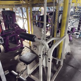 A seated leg curl machine with a padded seat and leg rollers is placed in a warehouse among other fitness equipment and metal shelving.