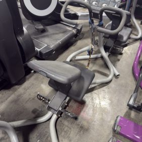 The Precor Stretch Trainer - As Is Functional, equipped with an instructional placard, is surrounded by various gym equipment on a concrete floor.