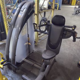 A seated chest press machine is positioned in an industrial warehouse setting with metal shelves and equipment visible in the background.