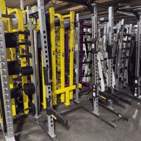 Several squat racks and weightlifting equipment are lined up in a gym, with weight plates stored on the sides and a concrete floor underneath.